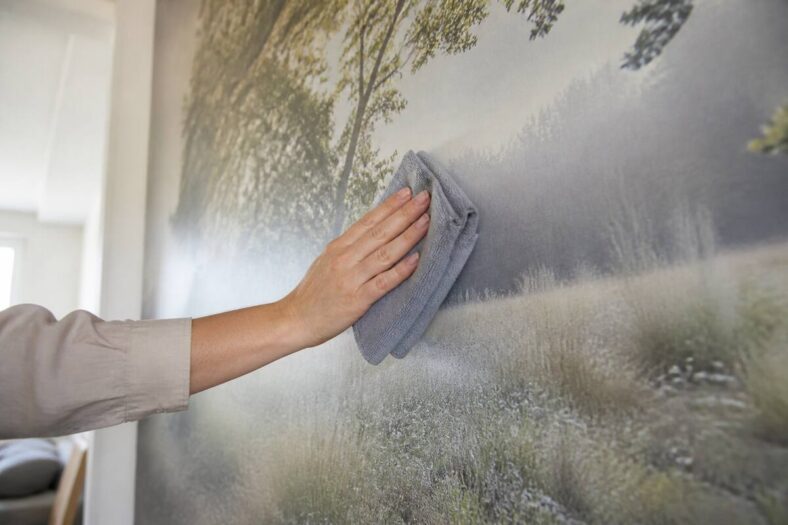 Iemand verwijdert voorzichtig stof van fotobehang met een zachte microvezeldoek in een lichte woonkamer